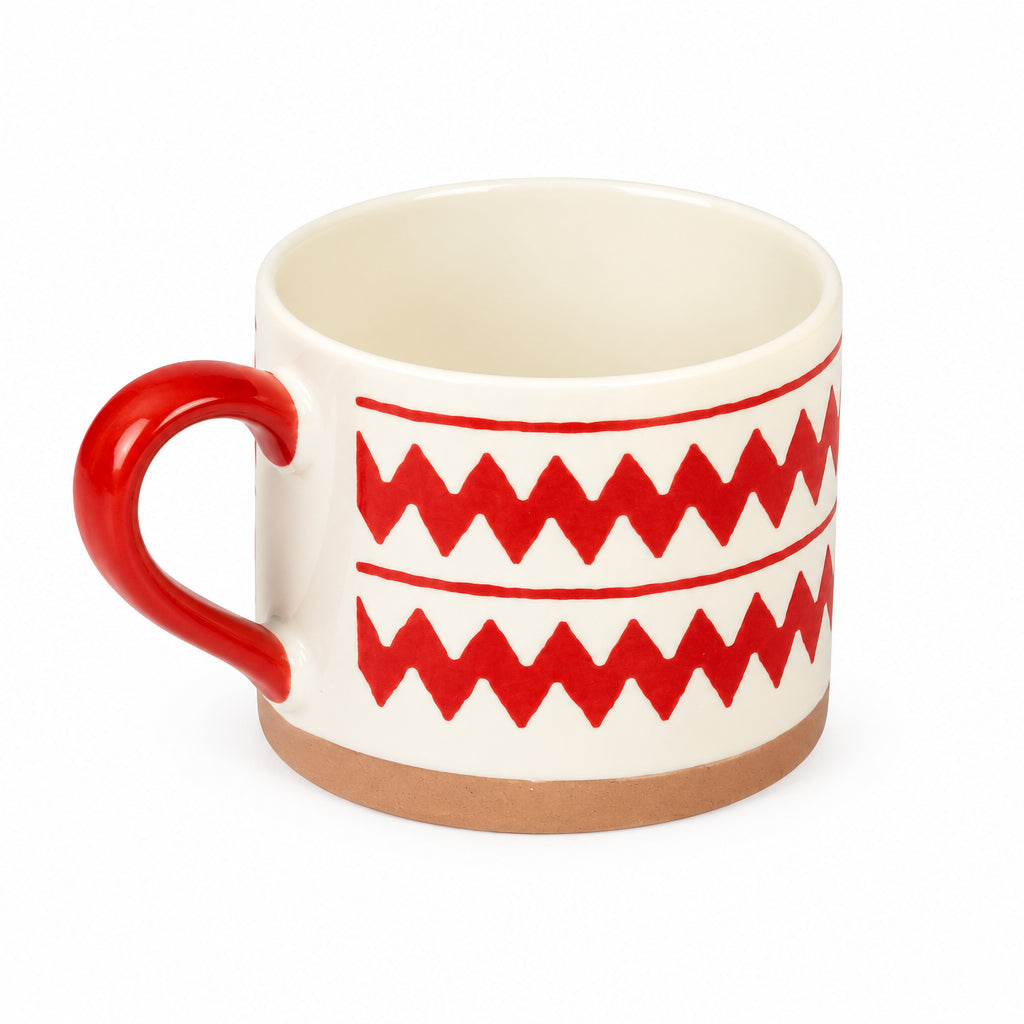 A ceramic mug with a cream-colored interior and exterior, decorated with a bold red zigzag pattern and a solid red handle, set against a plain light gray background.