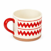 A ceramic mug with a cream-colored interior and exterior, decorated with a bold red zigzag pattern and a solid red handle, set against a plain light gray background.