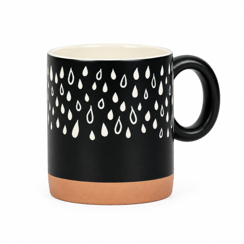 A ceramic mug with a matte black exterior decorated with a white raindrop pattern on the upper half and a terracotta-colored base, featuring a cream-colored interior and a black handle.