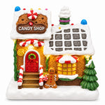 Load image into Gallery viewer, A small gingerbread-style candy shop figurine with a snow-covered roof, red door, decorated windows, a gingerbread man, and a small tree, photographed against a dark starry background.
